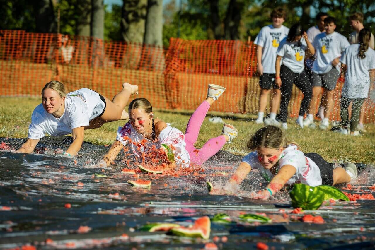 People sliding on watermelon-covered tarp outdoors.