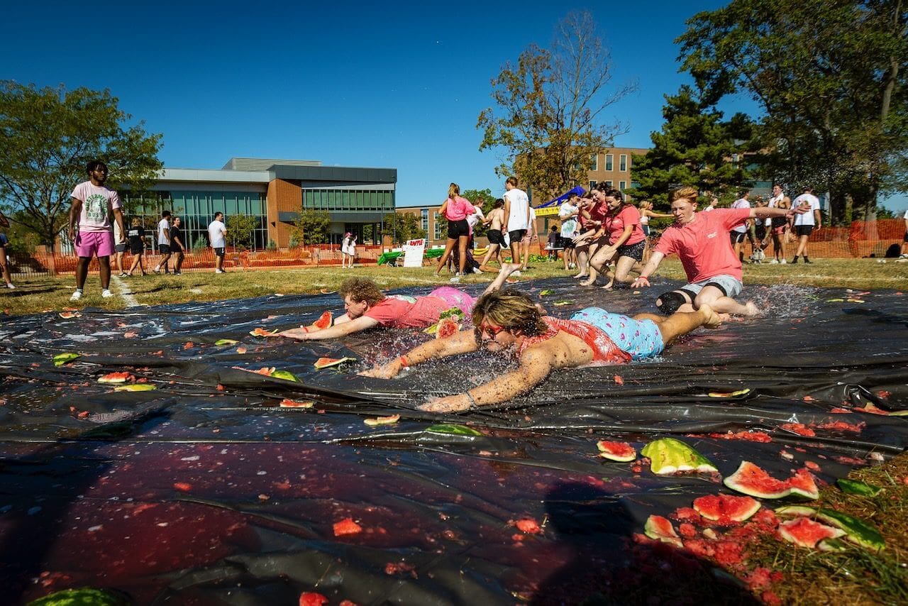 People sliding on watermelon-covered tarp outdoors.